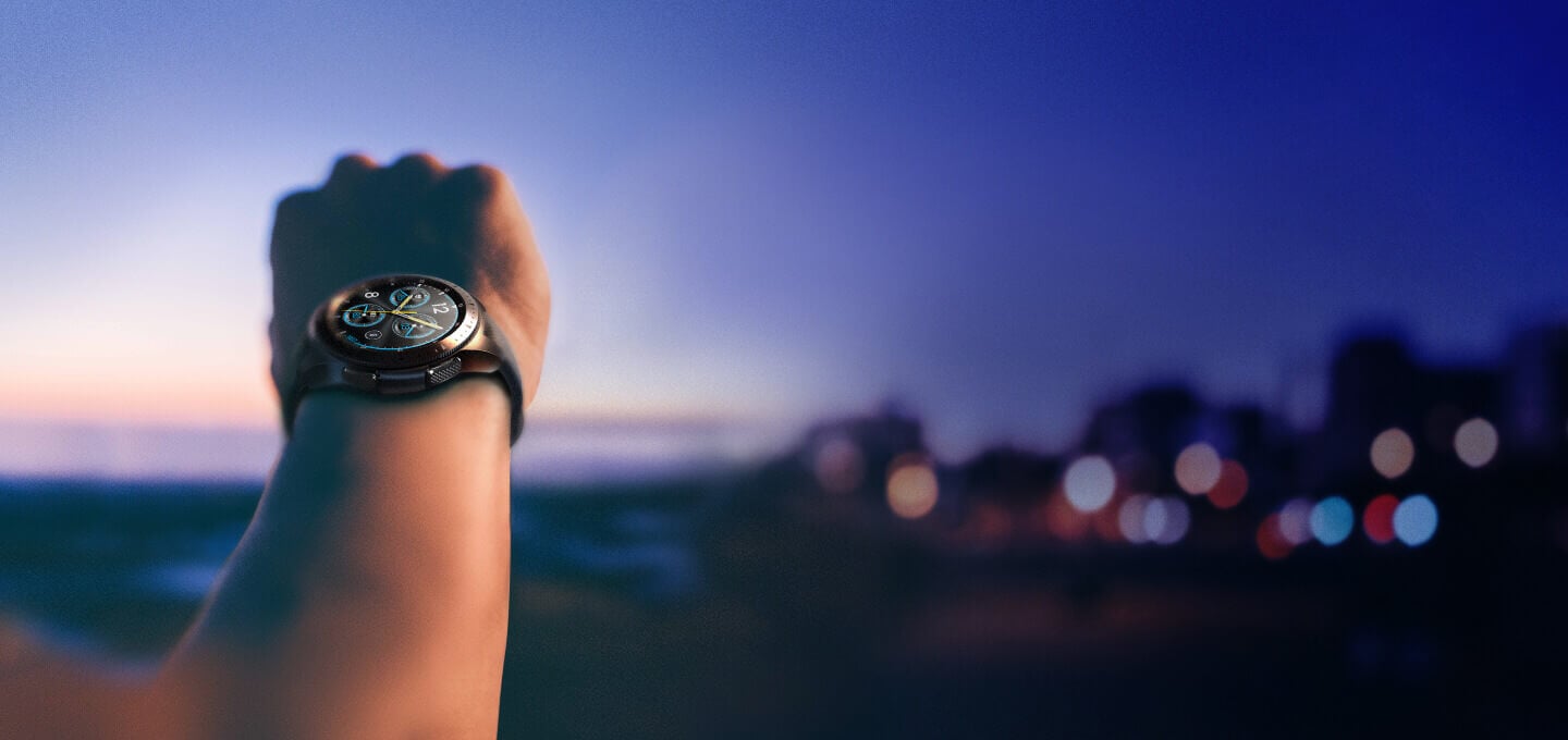A 42 mm Midnight Black Galaxy Watch on an unknown person's wrist with a blurred sunset and city view in the background.